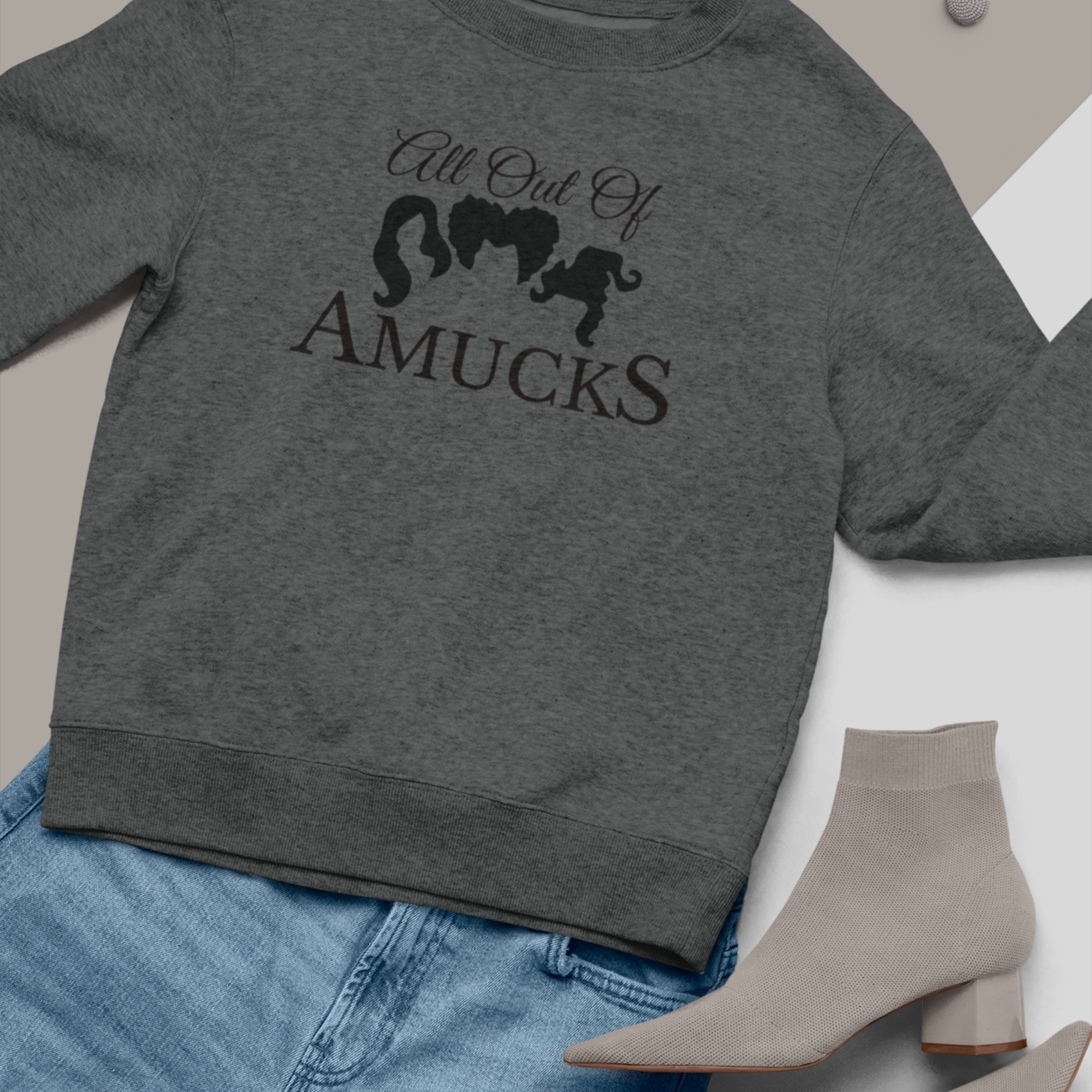 Dark gray cozy sweatshirt with the phrase "All Out of Amucks" featuring silhouettes of three witches' hairstyles, styled alongside light gray socks and blue jeans, perfect for Halloween or casual fall wear.