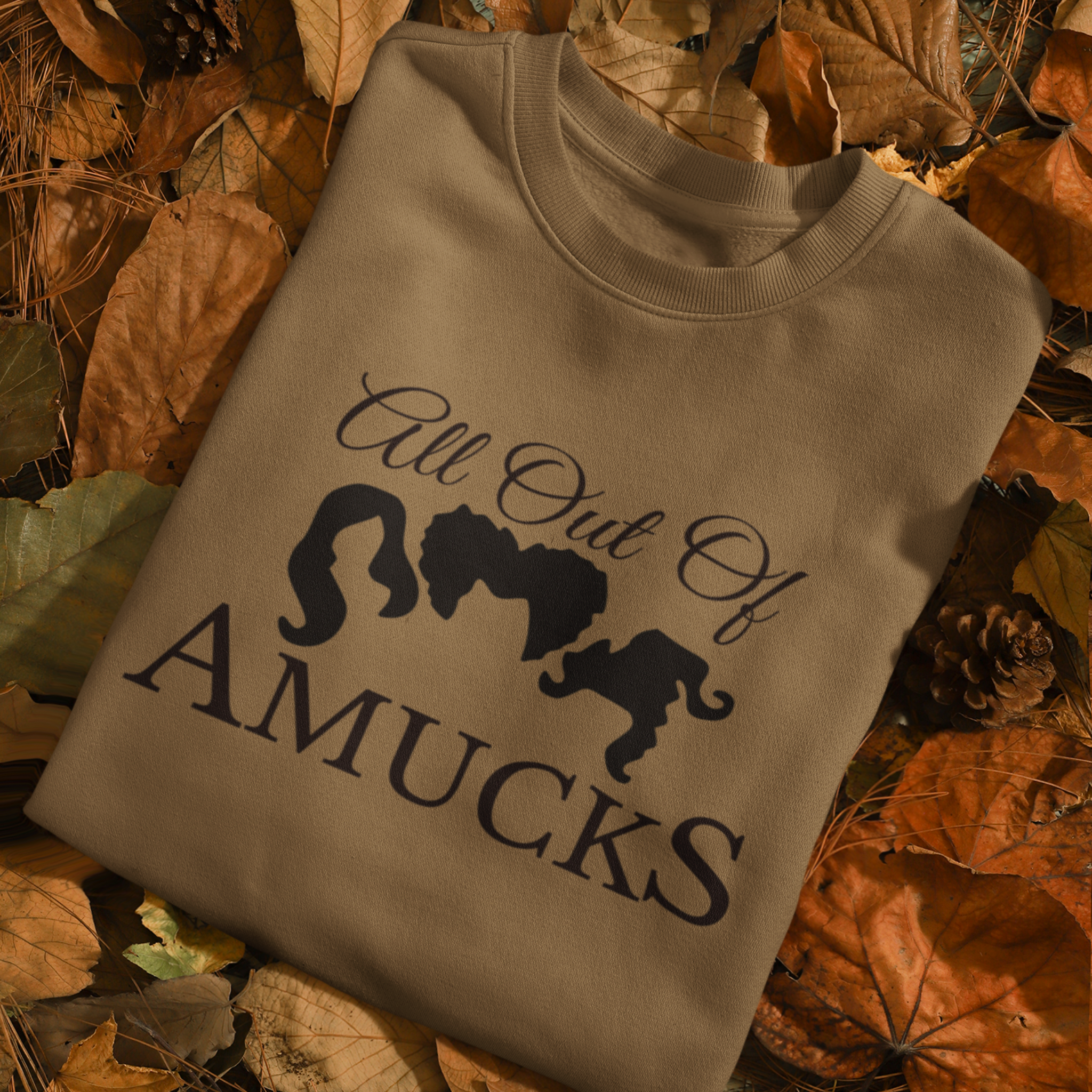 Folded brown sweatshirt with the phrase "All Out of Amucks" featuring silhouettes of three witches' hairstyles, placed on a bed of autumn leaves and pinecones, perfect for Halloween or fall-themed apparel.