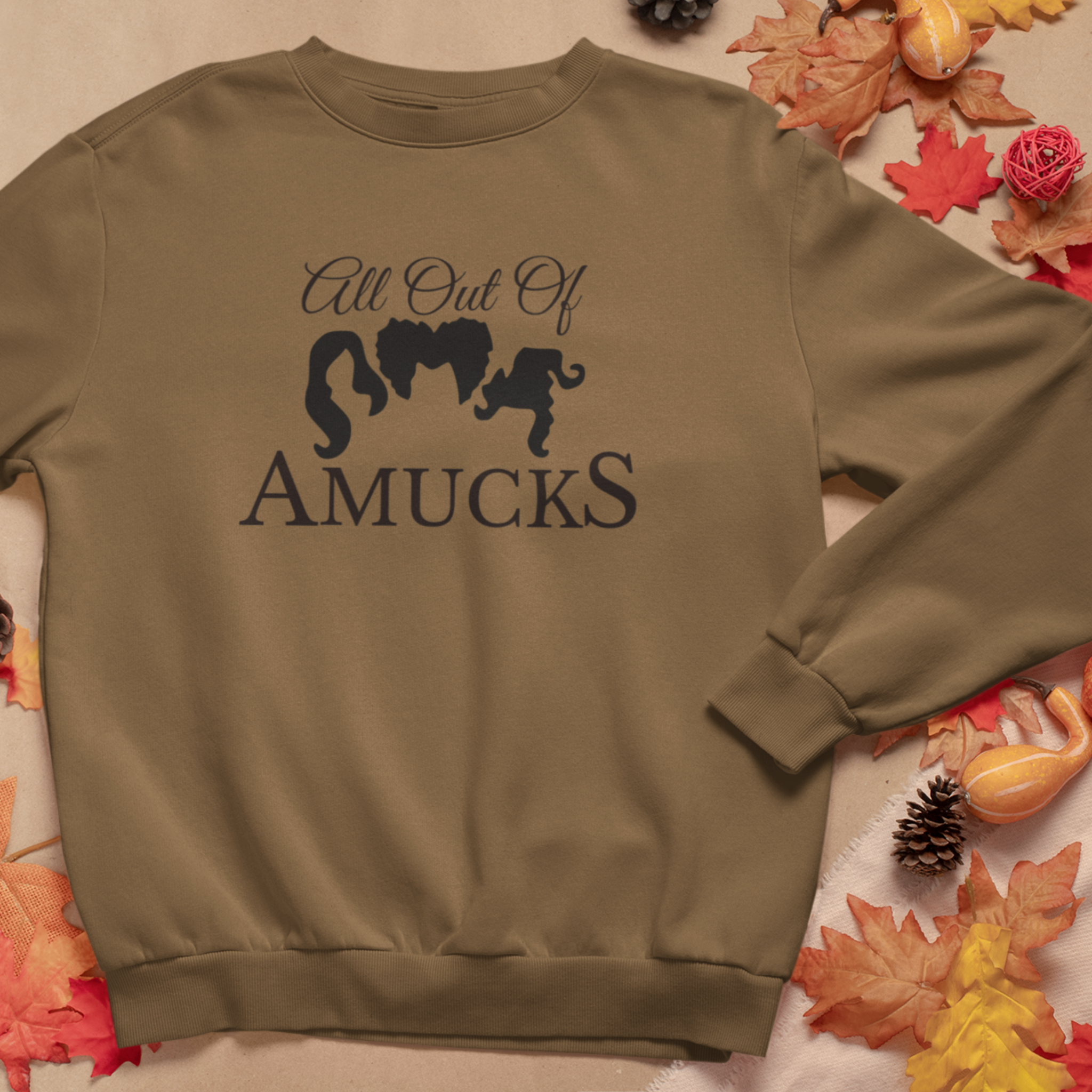 Brown cozy sweatshirt with the phrase "All Out of Amucks" featuring silhouettes of three witches' hairstyles, surrounded by fall leaves and pinecones, perfect for Halloween or autumn-themed outfits.