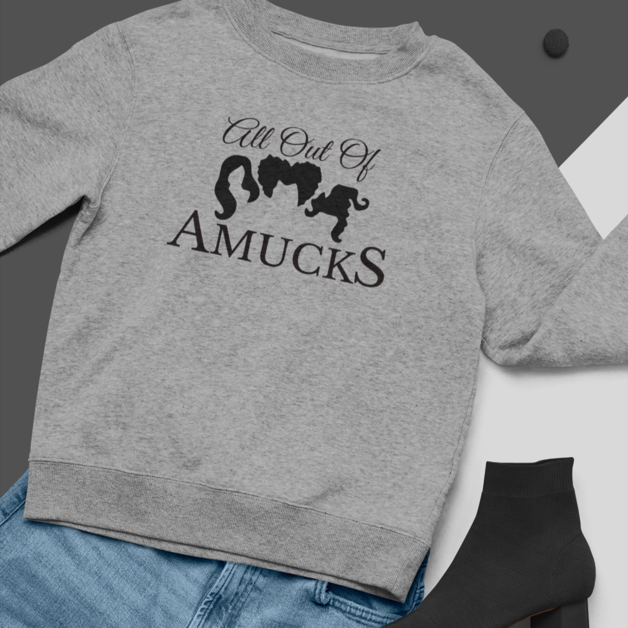 Gray cozy sweatshirt with the phrase "All Out of Amucks" featuring silhouettes of three witches' hairstyles, styled with blue jeans and black socks, perfect for Halloween or casual fall wear.