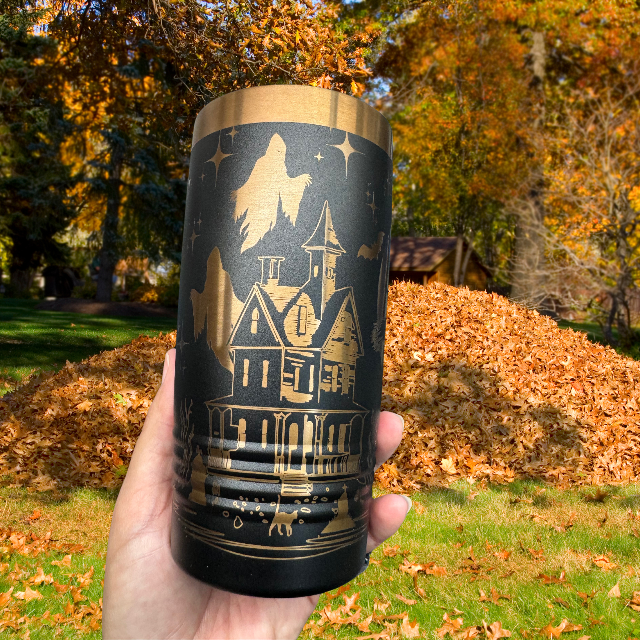 Black 20 oz laser-etched tumbler featuring a gold haunted house design with ghosts and bats, held outdoors in front of an autumn scene with fallen leaves and trees. Perfect for Halloween-themed drinkware.