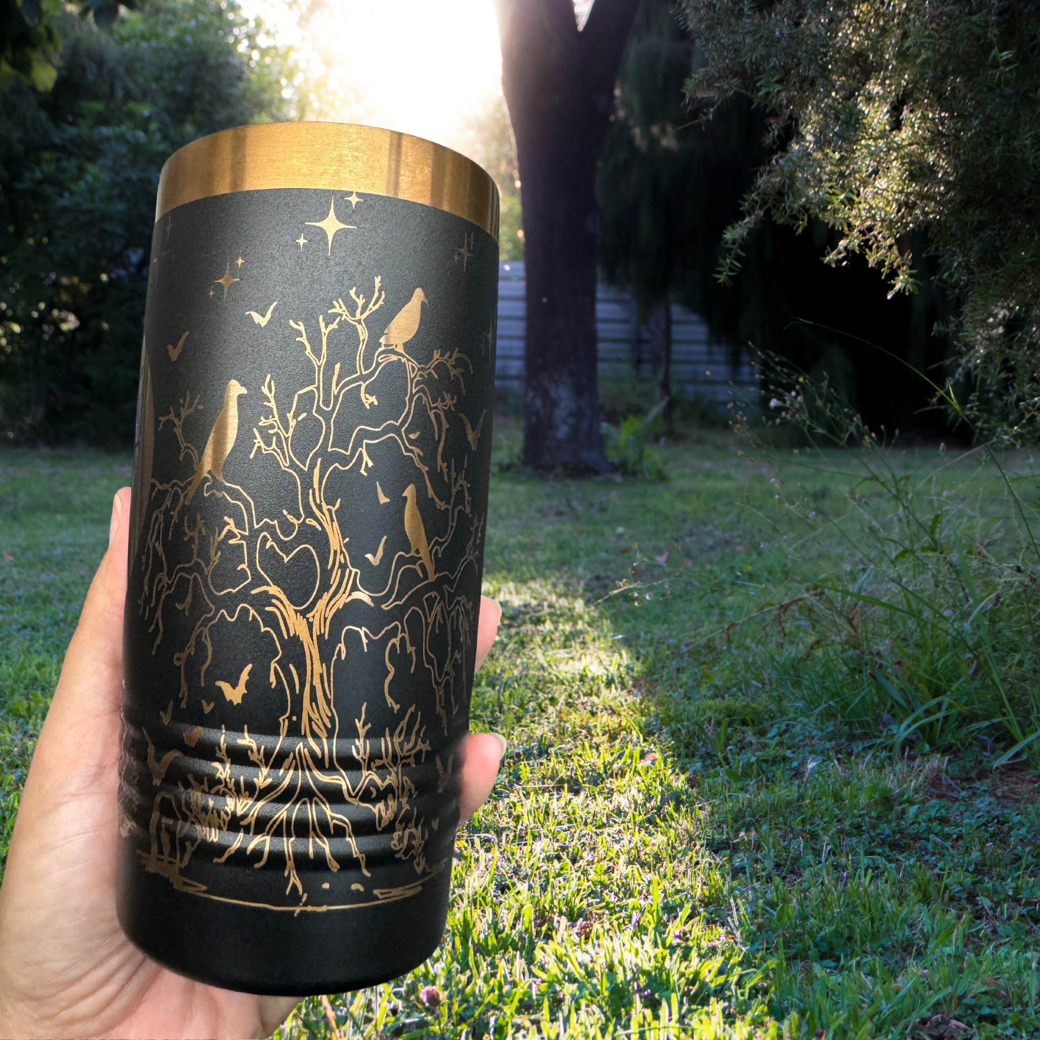 Black 20 oz laser-etched tumbler with a gold spooky tree design featuring ravens, bats, and stars, held outdoors in a grassy backyard with sunlight filtering through trees. Ideal for Halloween or gothic-themed drinkware.