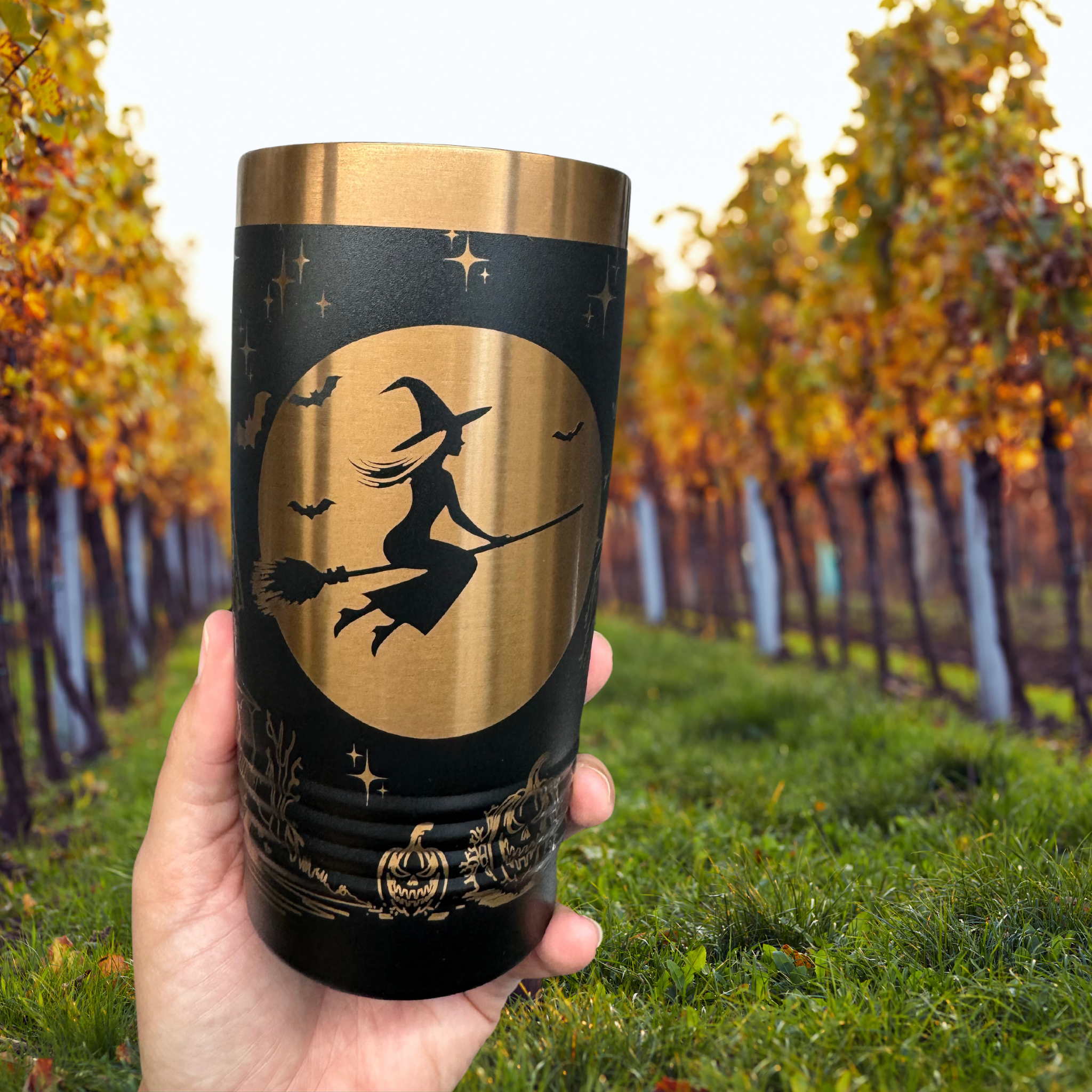 Black 20 oz laser-etched tumbler with a gold witch design flying on a broomstick against a full moon, held in front of a scenic vineyard with autumn-colored trees. Perfect for Halloween-themed drinkware.