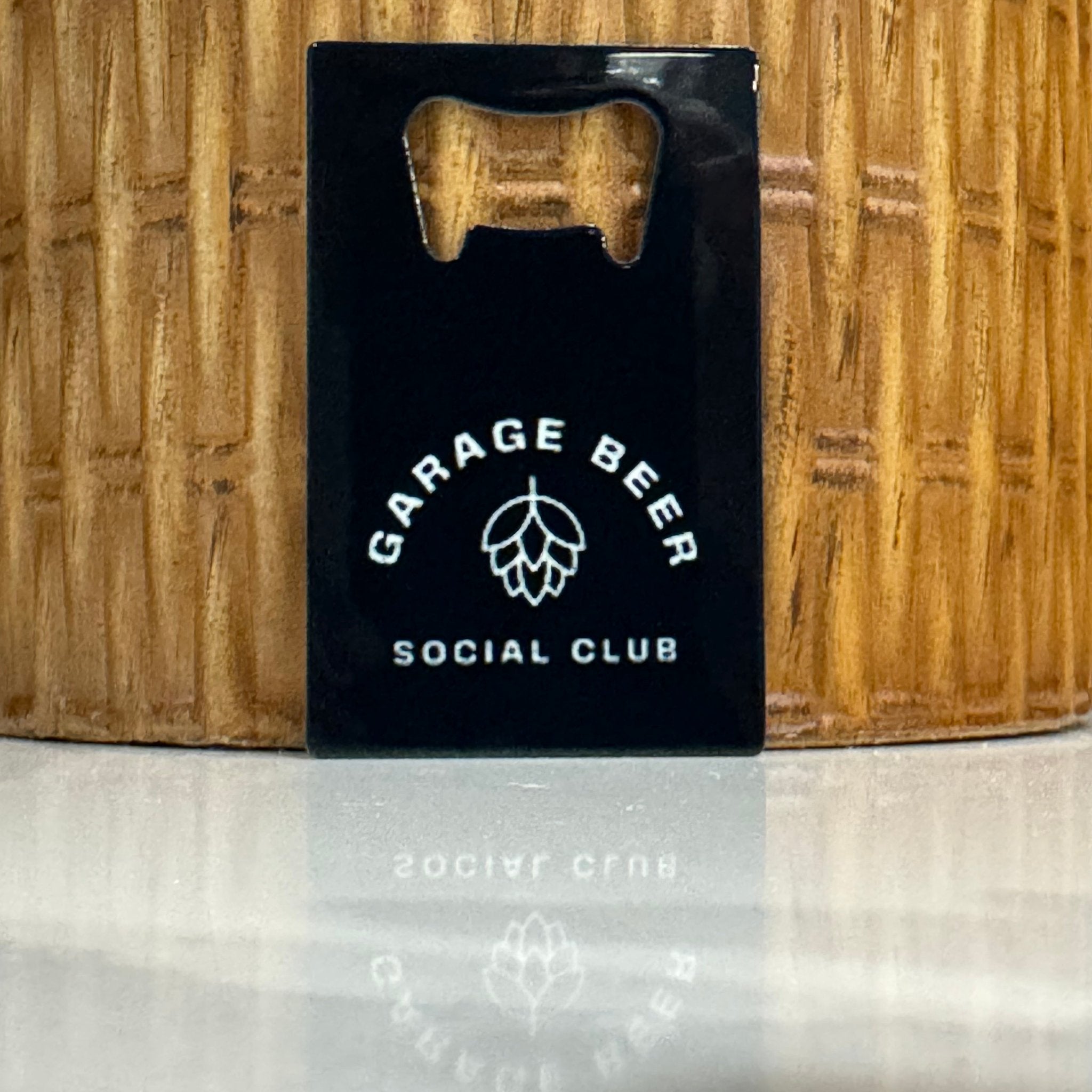 Black custom bottle opener business card with "Garage Beer Social Club" logo featuring a hop icon, positioned against a wicker background, ideal for craft beer branding and promotional items.