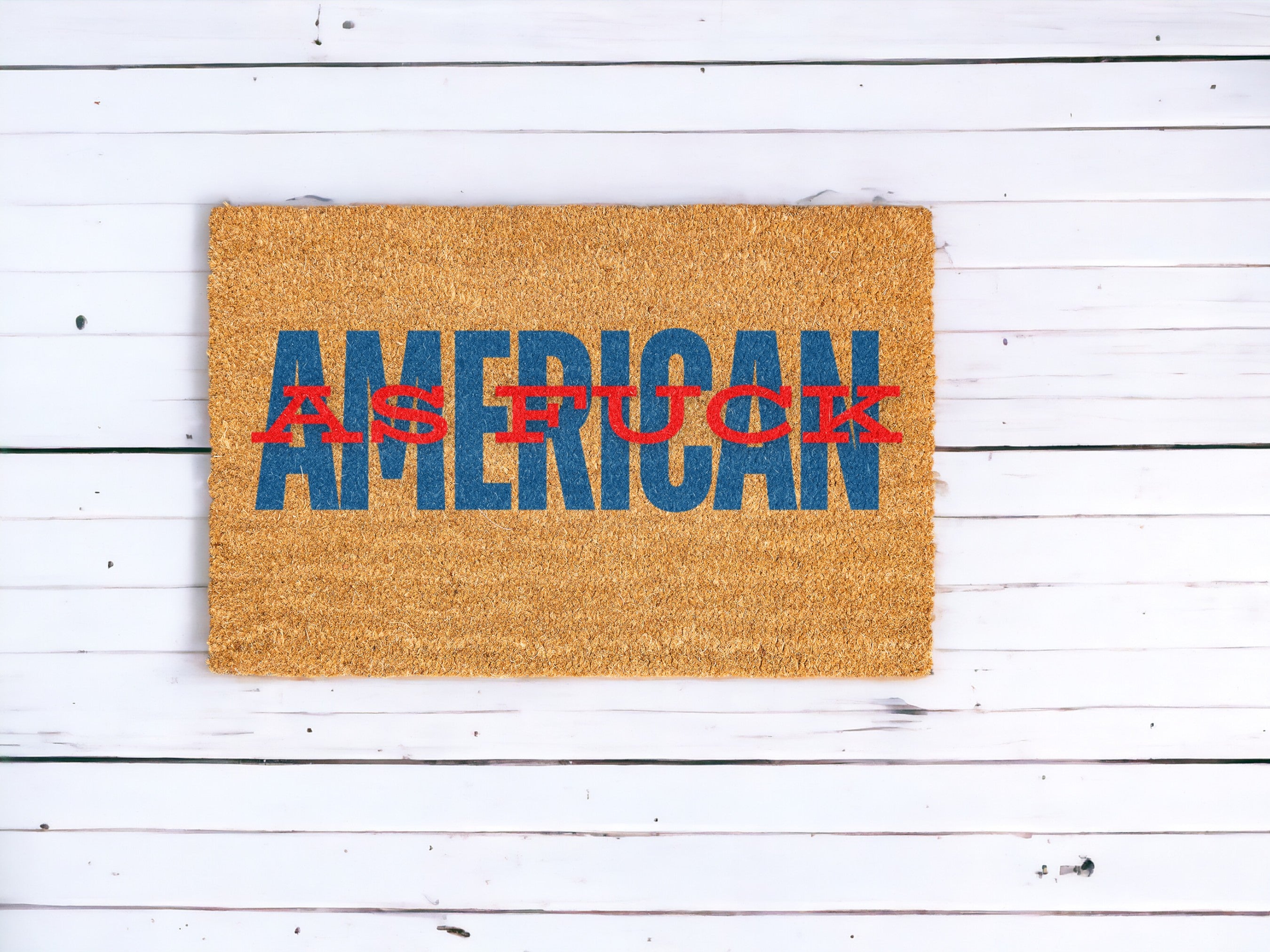 A brown, patriotic 'American As F*ck' Funny Doormat with a coarse texture displays the phrase "AMERICAN AS FUCK" in bold letters. The word "AMERICAN" is in large blue text, while "AS FUCK" is overlayed in red. This perfect Memorial Day decor sits on a white wooden floor with visible gaps between the planks.