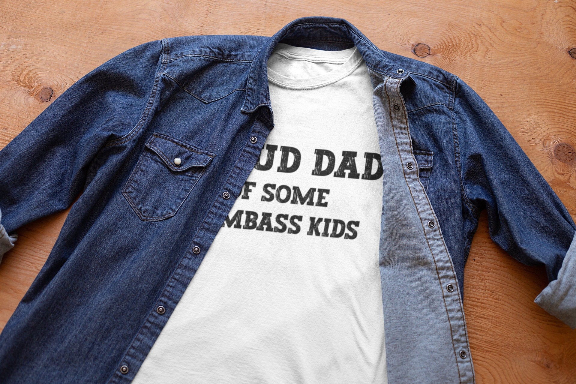 A white shirt under a jean jacket with the phrase 'Proud Dad of Some Dumbass Kids,' making it a humorous and custom Father's Day gift for dads. Ideal for those seeking unique and funny dad shirts.