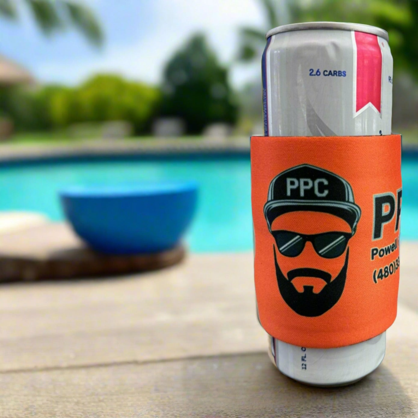 A can wrapped in an orange PPC-branded slap wrap drink sleeve sits on a poolside table, and a clear blue sky in the background. Ideal for promoting custom beverage sleeves in a tropical or resort setting.