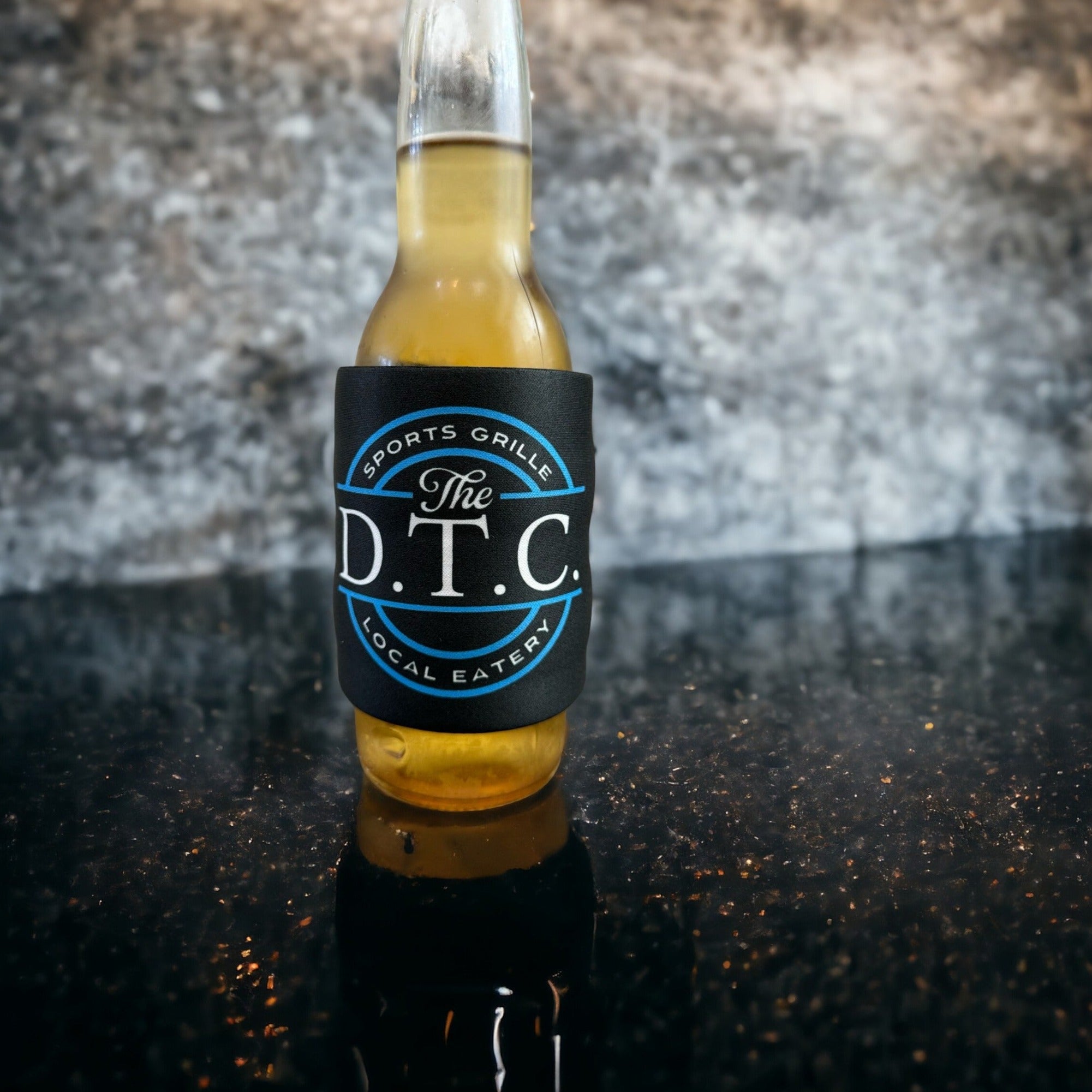 Glass bottle wrapped in a black slap wrap drink sleeve featuring "The D.T.C. Sports Grille Local Eatery" logo, placed on a reflective black surface, ideal for custom beverage holders and promotional items.